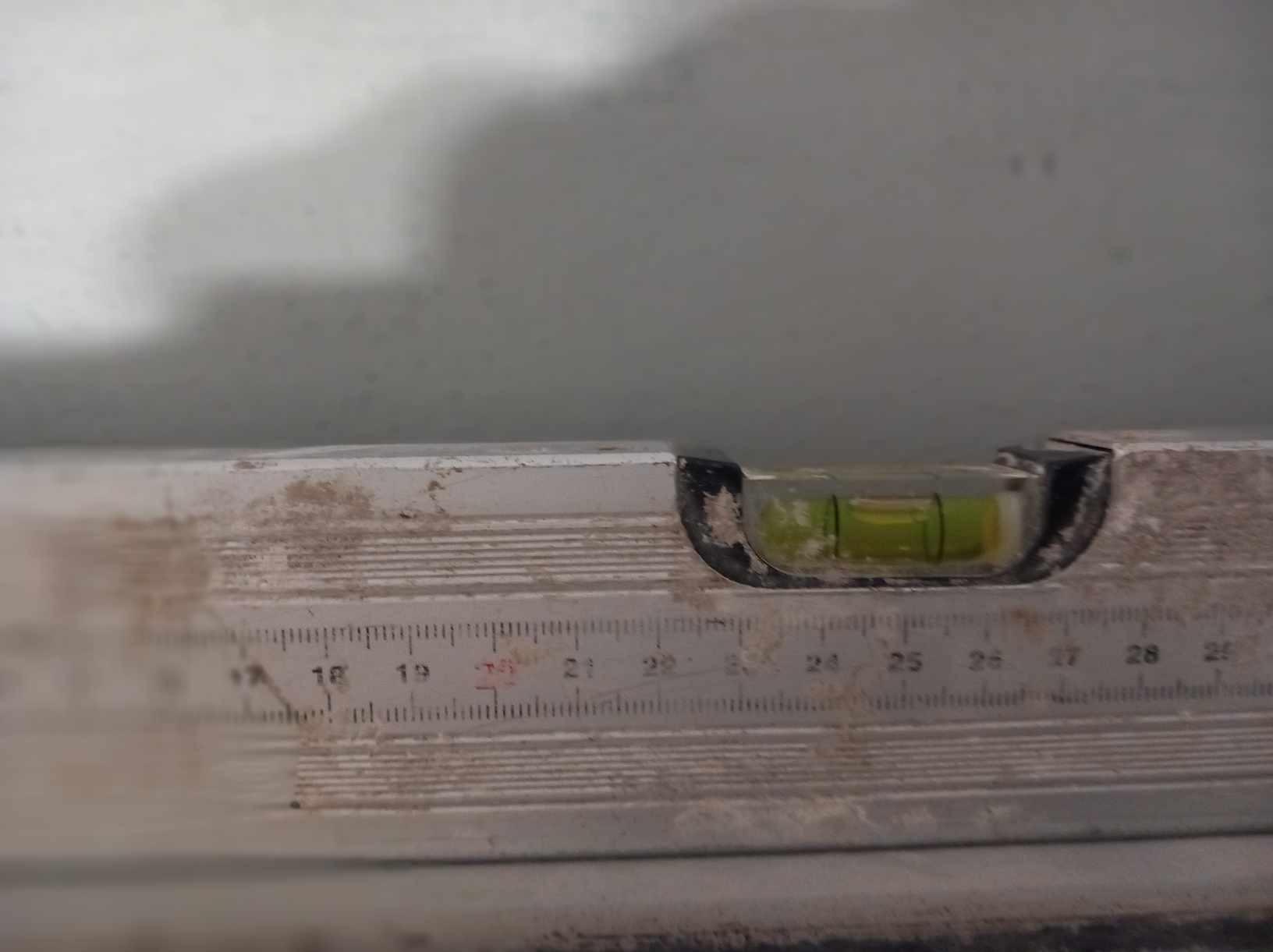 Poziomica z widoczną libelką na tle szarej ściany, widoczne drobne zabrudzenia na urządzeniu.