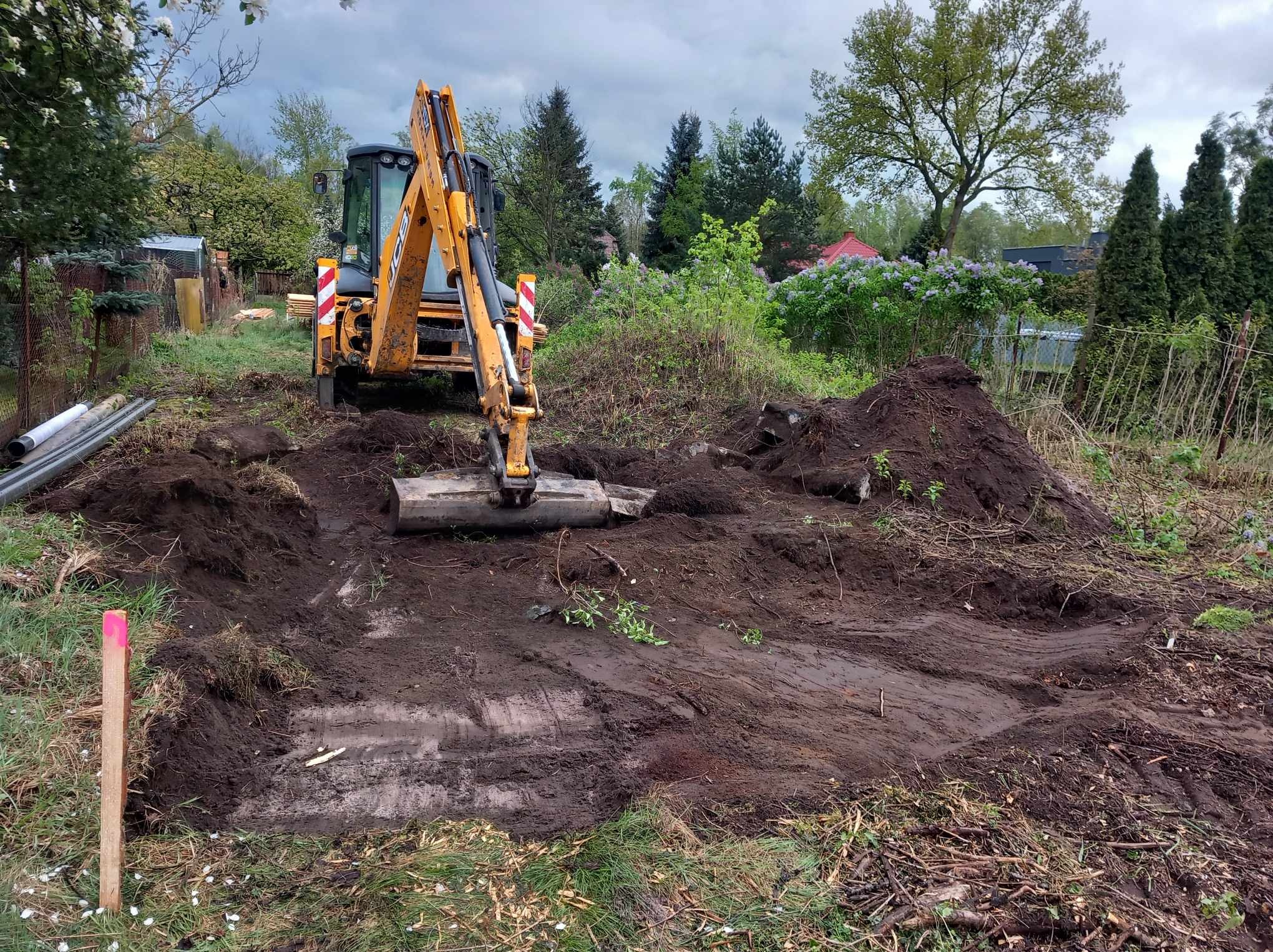 Żółta koparko-ładowarka na działce wyrównuje teren pod budowę, widoczne ślady opon i pryzma ziemi w tle, drewniany kołek z różową farbą w lewym dolnym rogu.