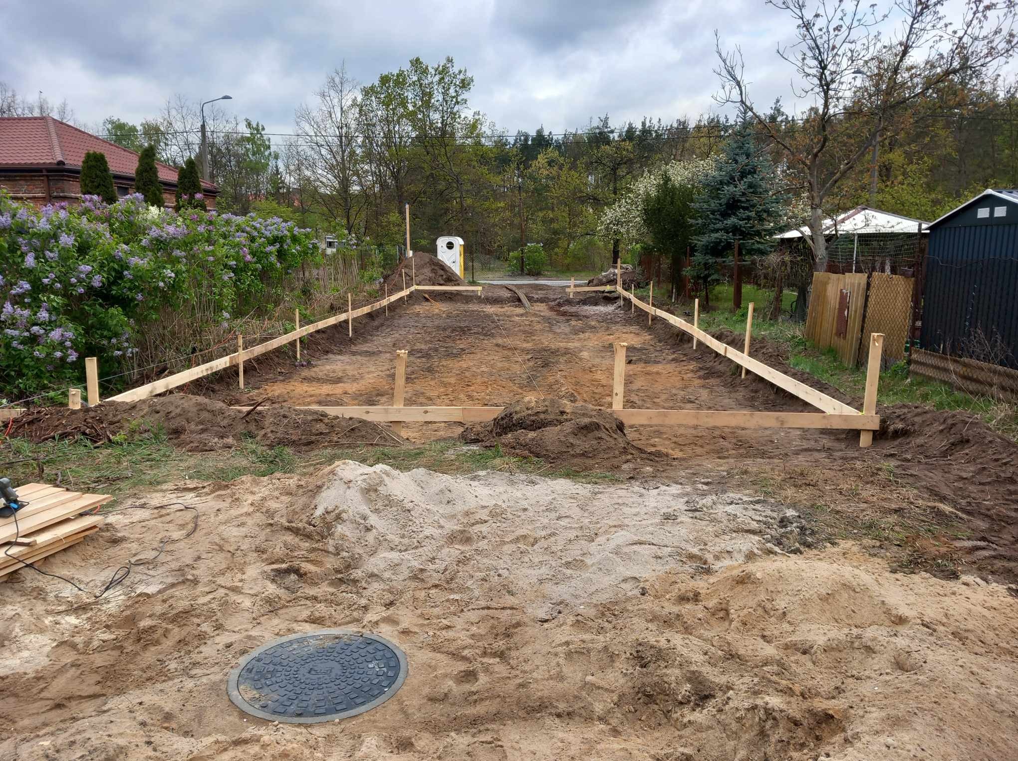 Wytyczony teren pod budowę domu z drewnianym szalunkiem, widoczny właz kanalizacyjny i tymczasowa toaleta przenośna.