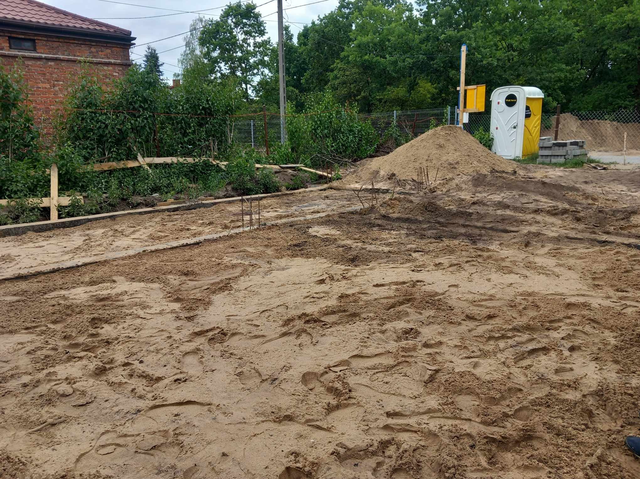 Przygotowany teren pod budowę z widocznymi śladami stóp na piasku, drewnianym szalunkiem i wystającymi prętami zbrojeniowymi, w tle pryzma piasku, przenośna toaleta i zieleń