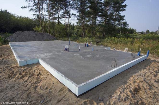 Wylana płyta betonowa z wystającymi rurami instalacyjnymi i prętami zbrojeniowymi, otoczona izolacją termiczną, na piaszczystym terenie budowy z widocznym kopcem ziemi w tle.