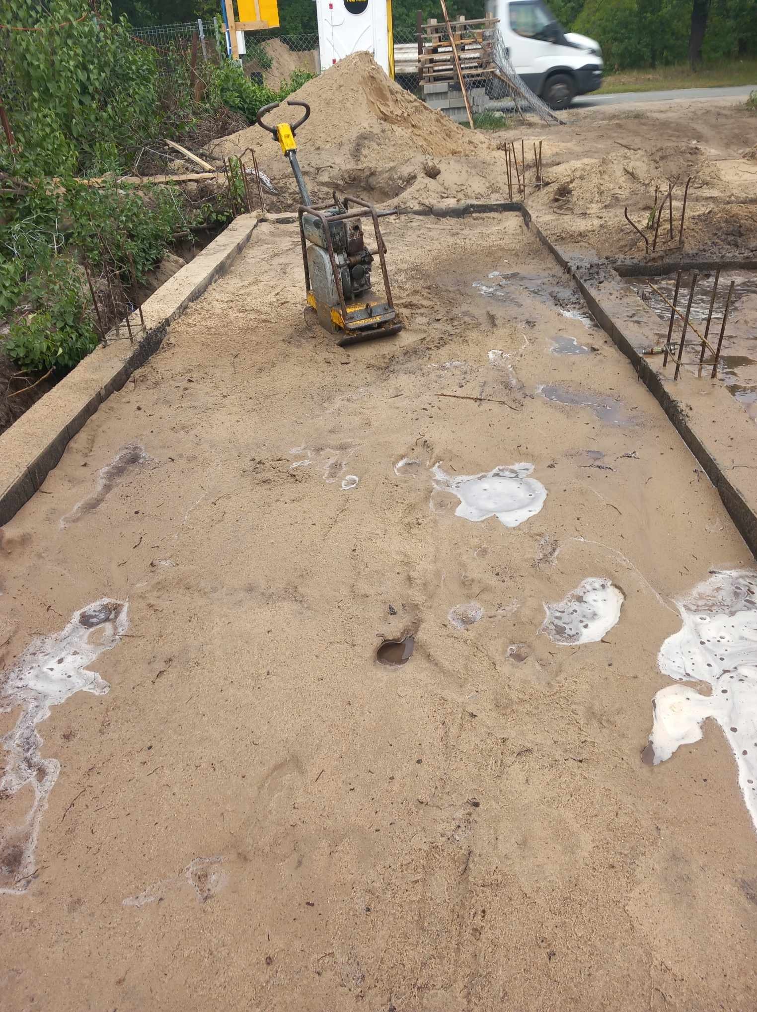 Wyrównany teren pod budowę z wystającymi prętami zbrojeniowymi i ubijarką wibracyjną na środku. Widoczne ślady wody i piany na piasku.