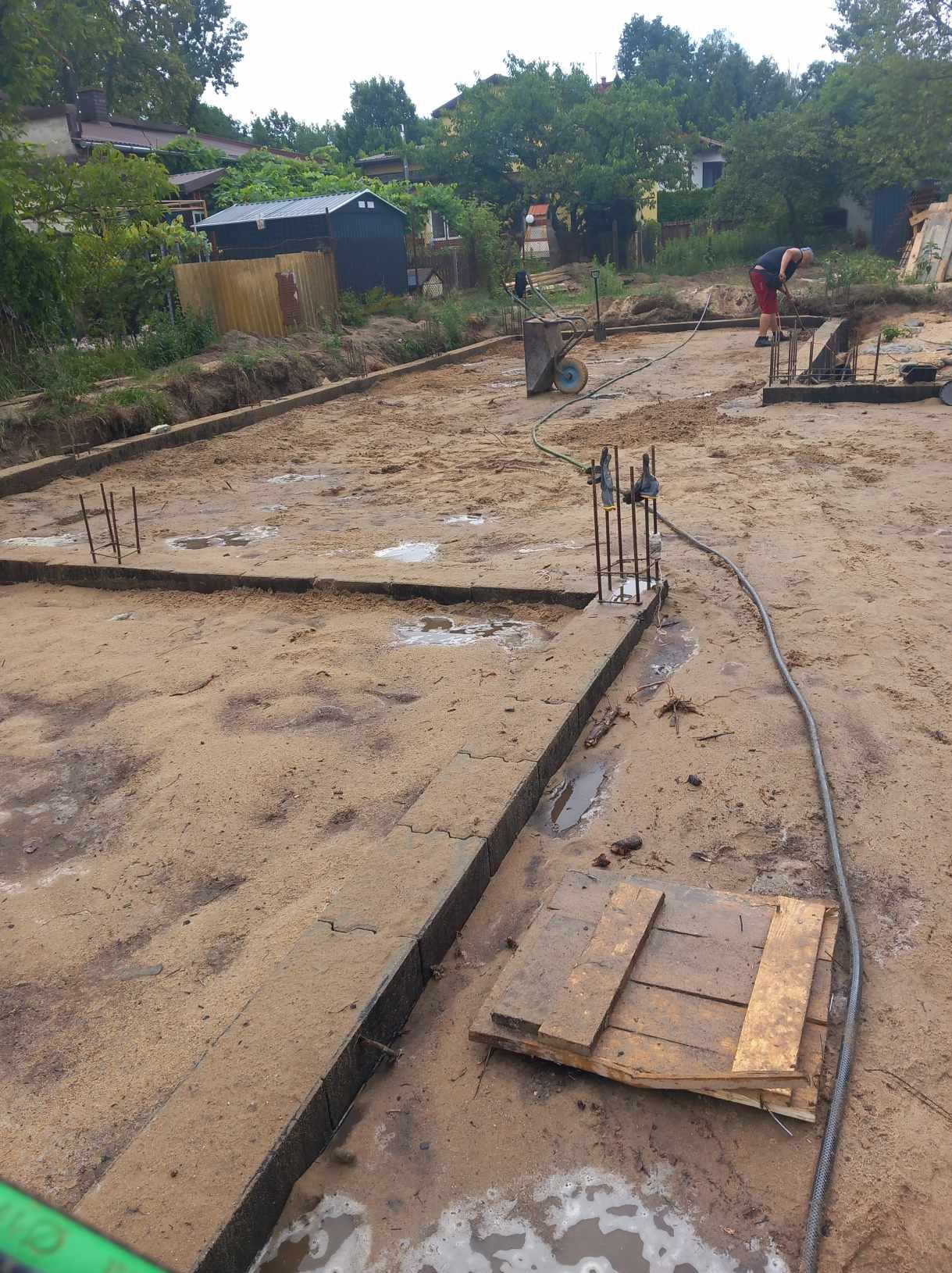 Przygotowanie terenu pod fundamenty: wykopane rowy, zbrojenie wystające z ziemi, pracownik w tle, widoczne ślady po deszczu i błoto.
