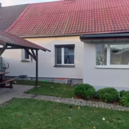 Jasna elewacja domu jednorodzinnego z czerwonym dachem, widok z ogrodu. Elementy architektury ogrodowej i roślinność w kadrze.