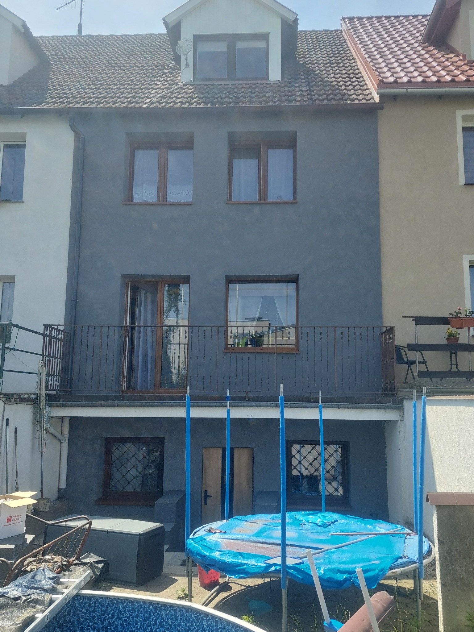 Dom po remoncie elewacji w odcieniach szarości, z widocznym balkonem i fragmentem trampoliny w ogrodzie. Nowe okna i świeża farba podkreślają efekt modernizacji.