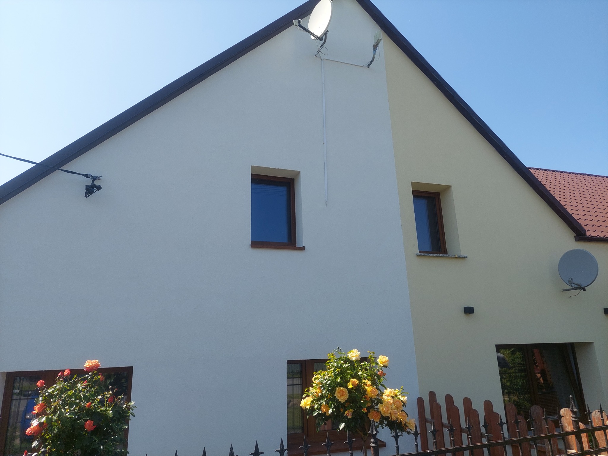 Dom jednorodzinny z ocieploną, dwukolorową elewacją, widoczne okna z brązowymi ramami, satelity na dachu i kwiaty przed ogrodzeniem.