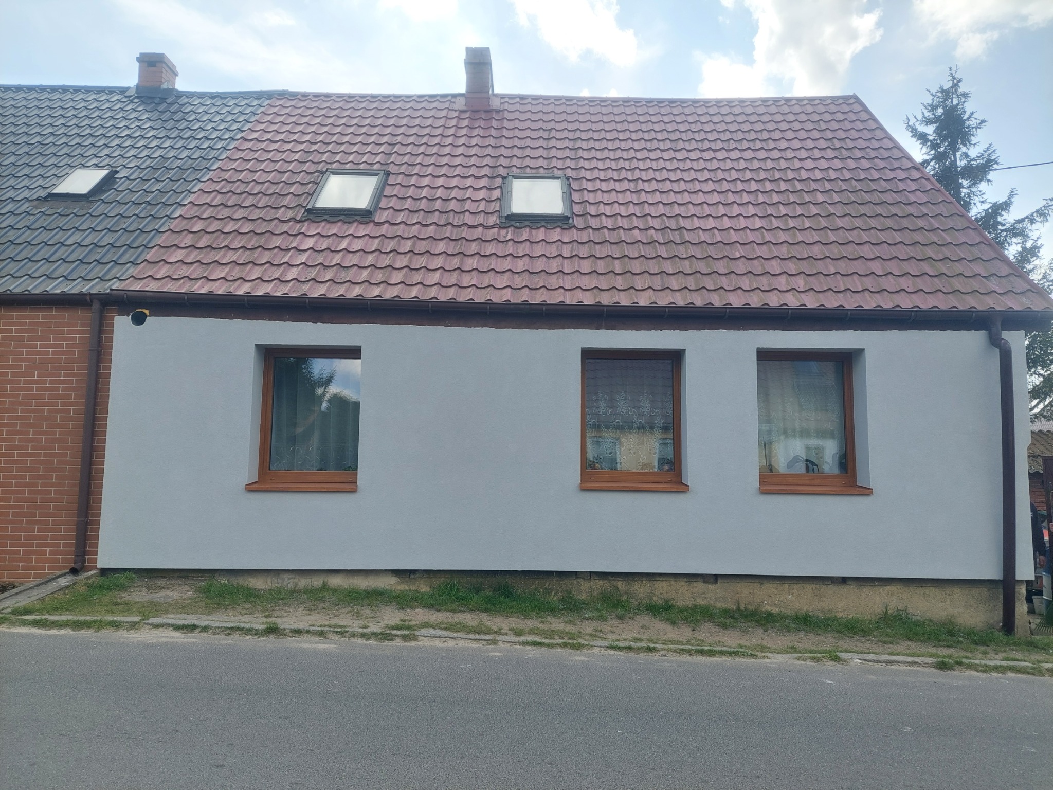Dom jednorodzinny po remoncie elewacji, z widocznymi trzema oknami z drewnianymi ramami i dachem z czerwonej dachówki. Część dachu pokryta czarną dachówką.