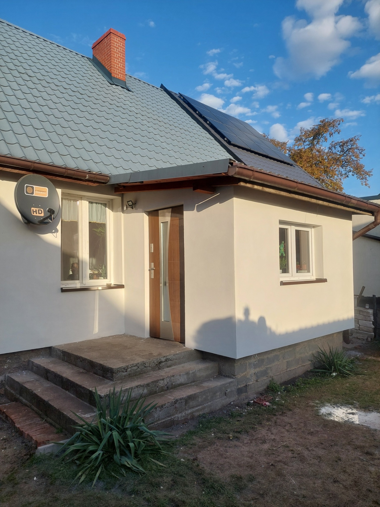 Biała elewacja domu jednorodzinnego z brązowymi akcentami, widoczne panele słoneczne na szarym dachu i antena satelitarna. Ujęcie w słoneczny dzień.