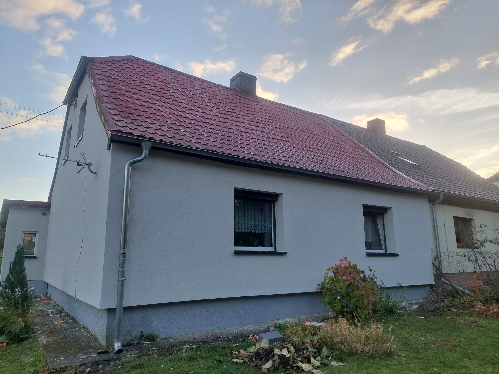 Świeżo otynkowany dom z czerwoną dachówką i szarym cokołem, otoczony zielenią i błękitnym niebem. Widoczne okna z roletami i rynna. Elewacja po remoncie.