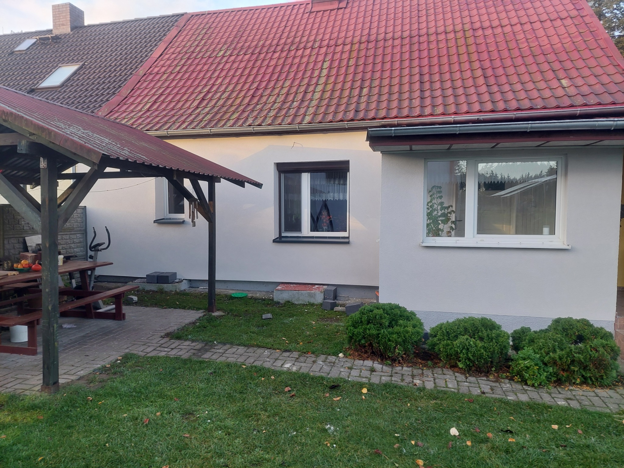 Jasna elewacja domu jednorodzinnego z czerwonym dachem, widok z ogrodu. Elementy architektury ogrodowej i roślinność w kadrze.