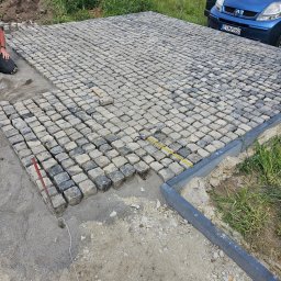 Pmbud - Fragment powierzchni z kostki granitowej w trakcie układania, widoczna część ułożona, piasek podsypkowy, obrzeża betonowe, narzędzia pracy, w tle trawa i zaparkowany niebieski samochód.