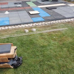 Pmbud - Pracownik układa kolorowe, gumowe płyty chodnikowe na trawniku, obok widoczne pakiety płyt i narzędzia.