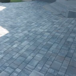 Nowo ułożona, szara kostka brukowa w jodełkę prowadząca do schodów wejściowych domu z kolumną, otoczona zielonym trawnikiem z ozdobnymi krzewami.