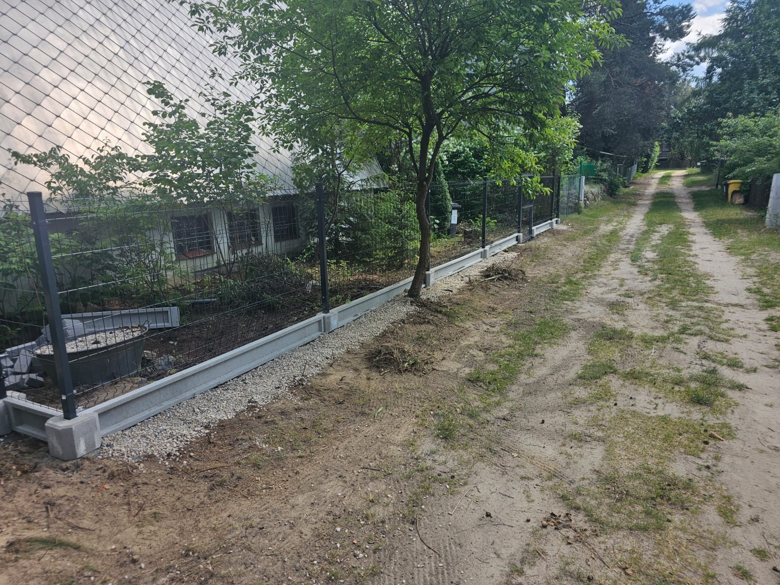 Montaż ogrodzenia panelowego z podmurówką betonową, widoczne elementy w trakcie instalacji, podsypka żwirowa, tło z budynkiem i drzewami.