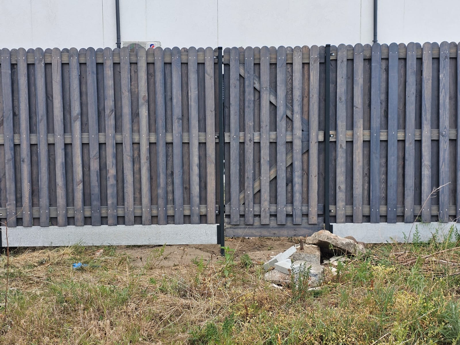 Drewniany płot panelowy pomalowany na szaro z bramką, osadzony na betonowej podmurówce, widoczne pozostałości gruzu i trawa.
