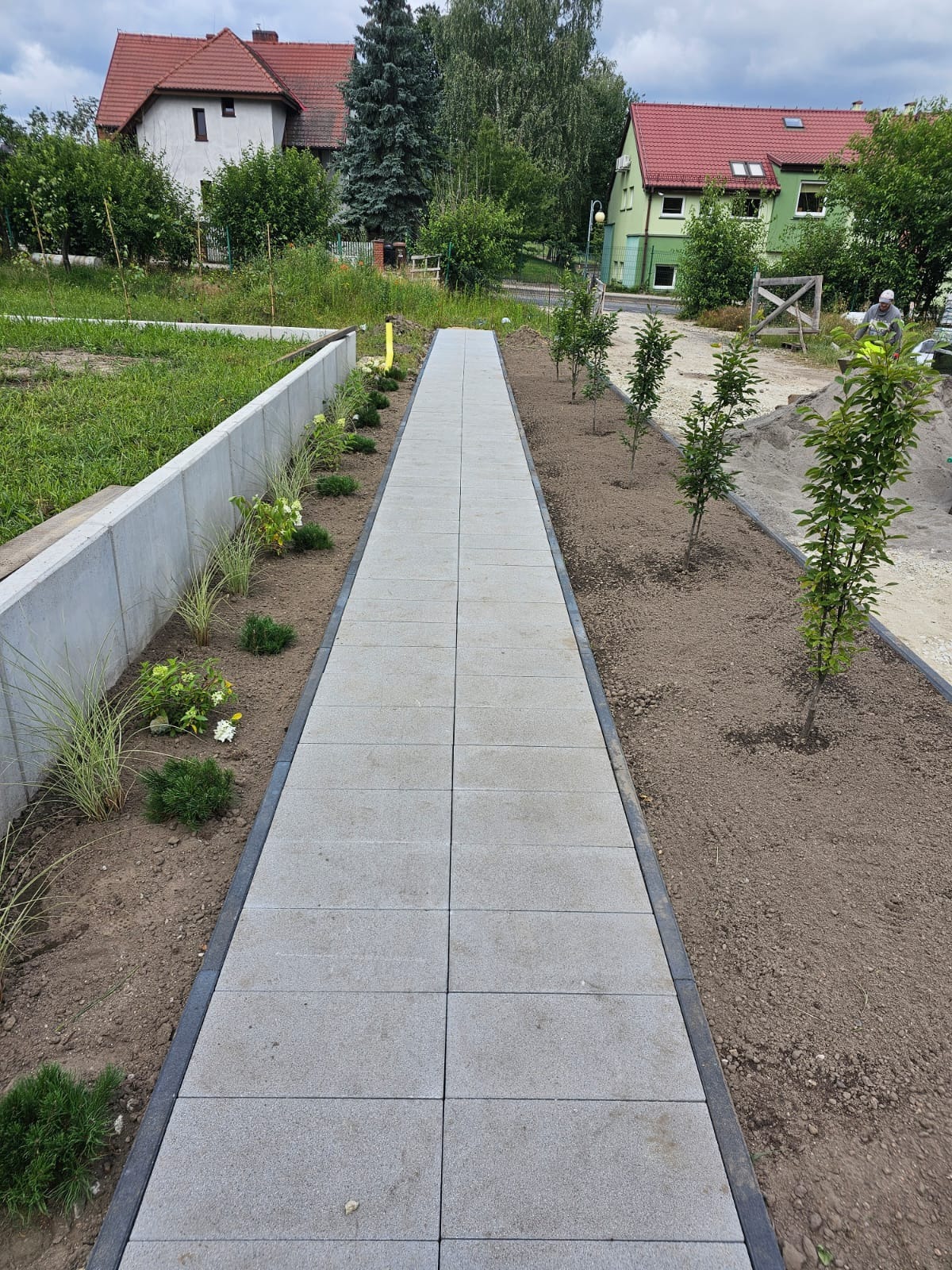 Nowa Sól: Prosta ścieżka z szarych płyt betonowych z czarnym obramowaniem, świeżo obsadzona roślinami i młodymi drzewkami po obu stronach, zakończona niskim betonowym murem.