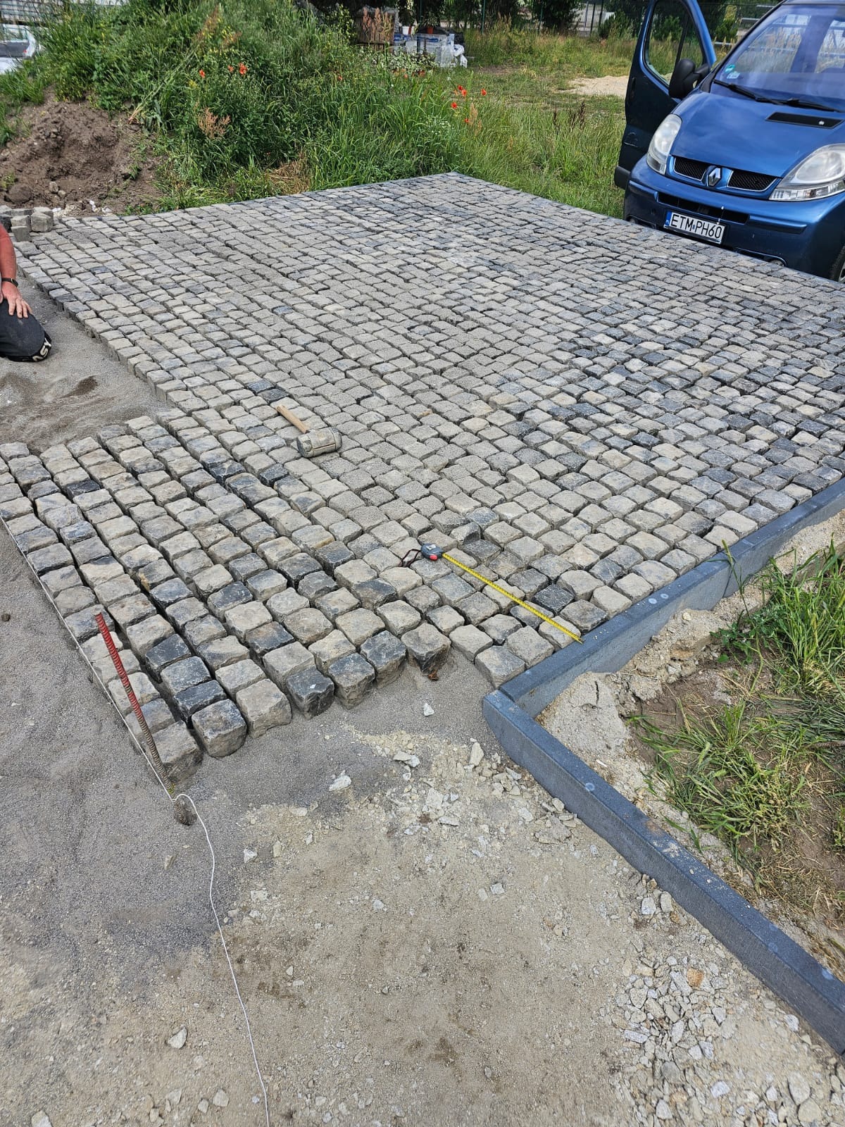 Fragment powierzchni z kostki granitowej w trakcie układania, widoczna część ułożona, piasek podsypkowy, obrzeża betonowe, narzędzia pracy, w tle trawa i zaparkowany niebieski samochód.