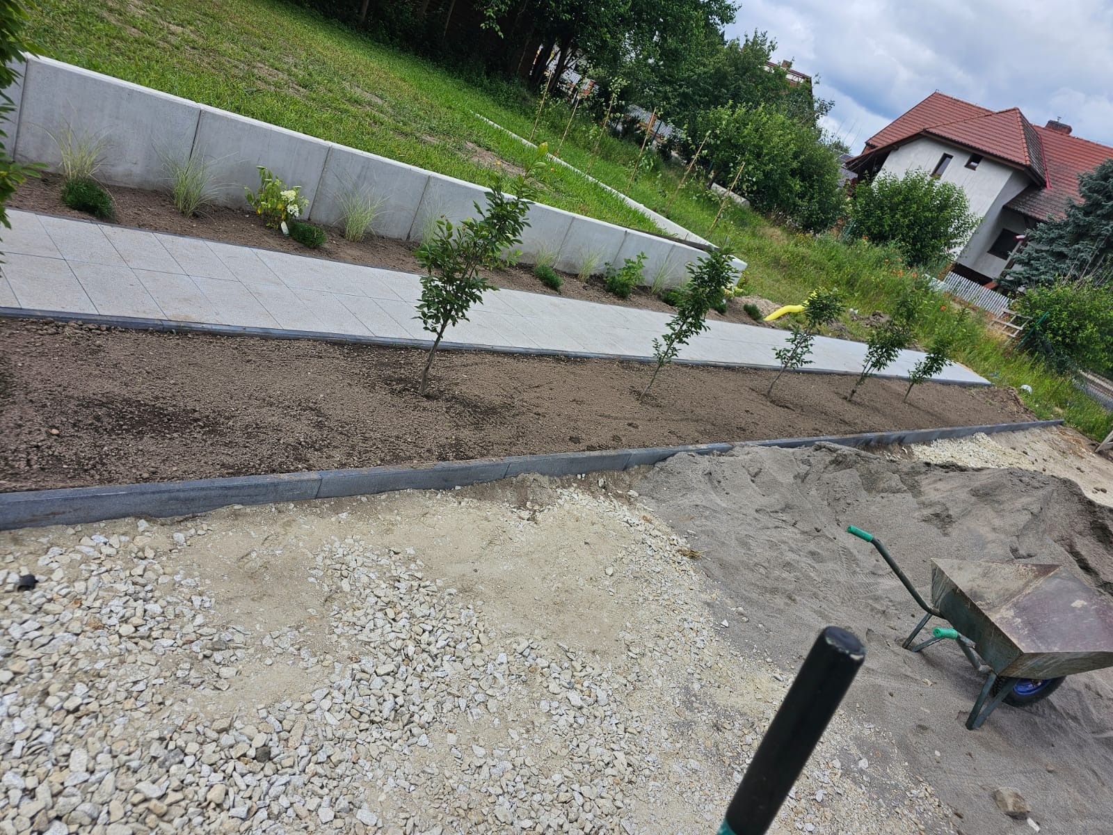 Ścieżka z jasnoszarych płyt prowadząca wzdłuż betonowego murku z nasadzeniami, obok świeżo posadzone drzewka, widoczny fragment terenu przygotowanego pod utwardzenie z piaskiem, żwirem i taczką.