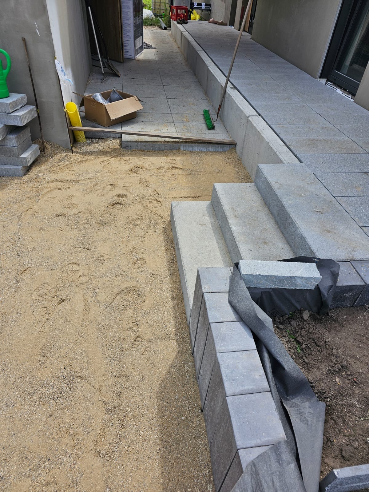 Przygotowanie podłoża pod kostkę brukową: warstwa piasku z widocznymi śladami stóp, ułożone schody z kostki, paleta kostki, narzędzia budowlane i fragment ułożonej ścieżki z kostki na zewnątrz...