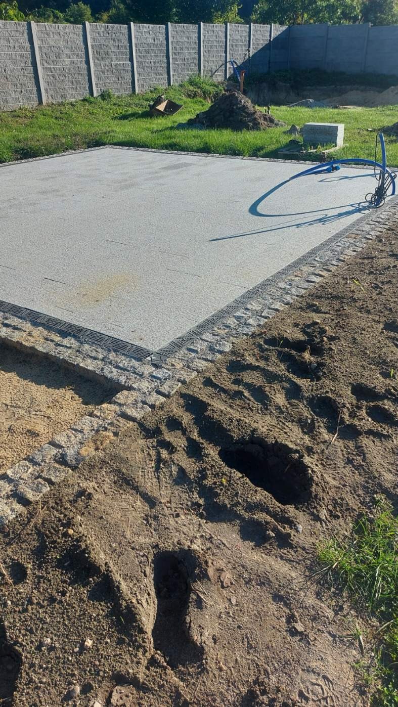 Przygotowany plac pod budowę z widoczną wylaną betonową płytą, obrzeżami wykończonymi kostką granitową i systemem odprowadzania wody, w tle betonowe ogrodzenie.