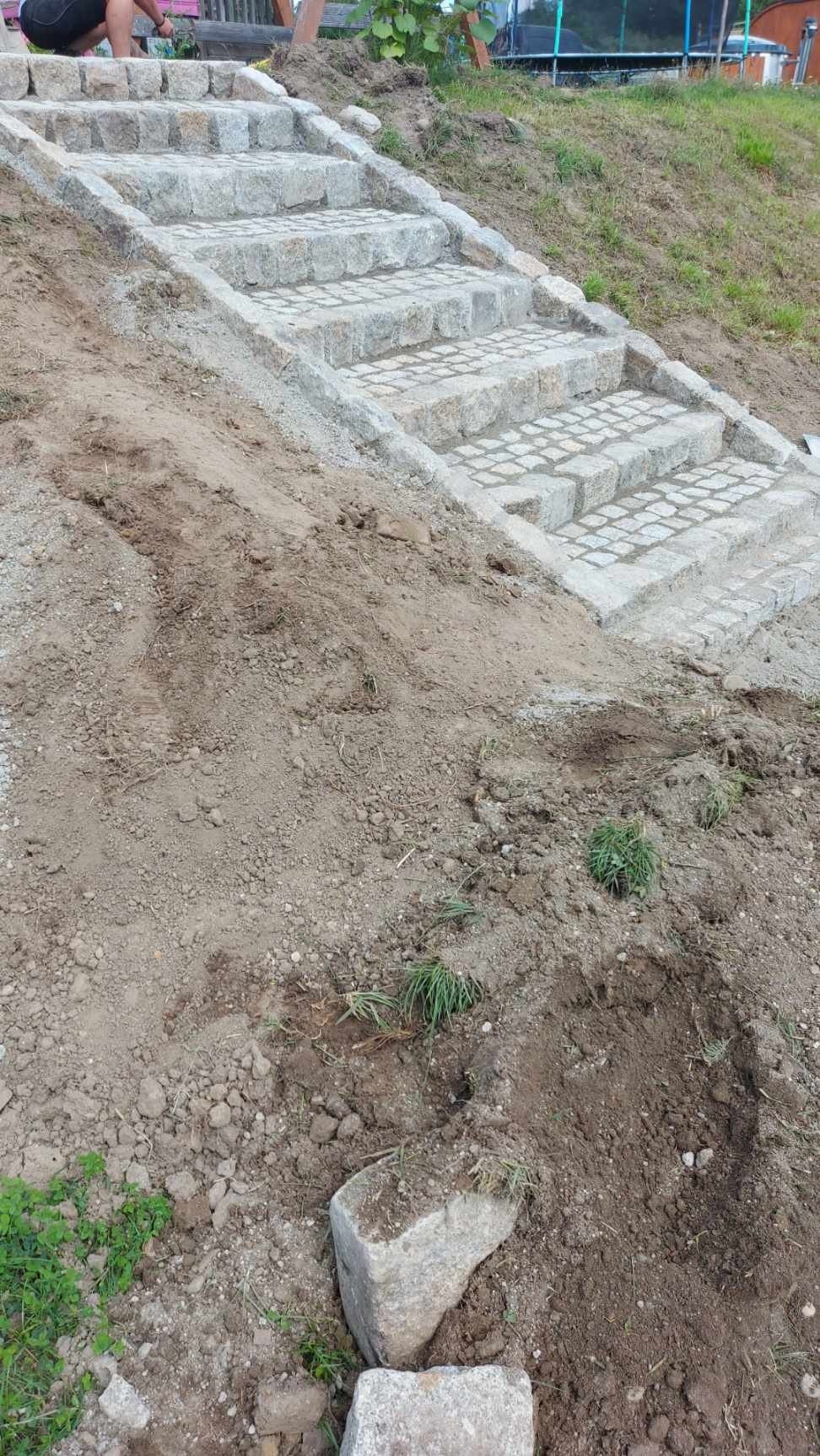 Kamienne schody ogrodowe prowadzące w górę zbocza, wykonane z jasnoszarej kostki granitowej, widoczne ślady prac ziemnych i niewielkie fragmenty trawy.