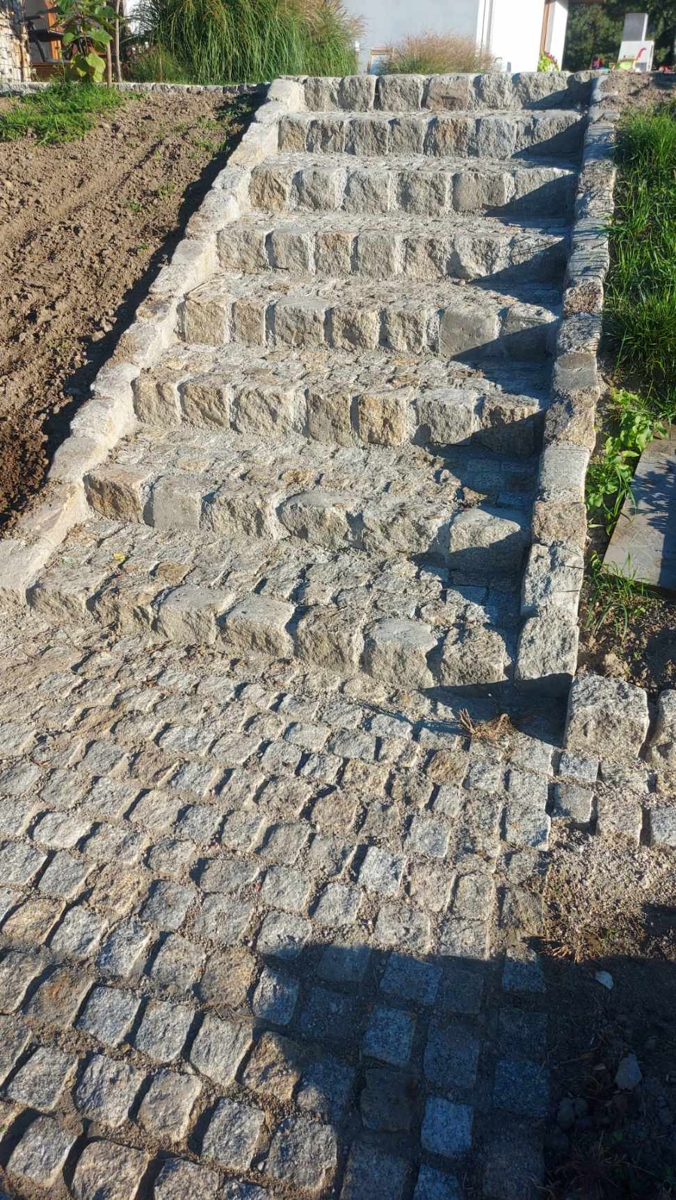 Schody zewnętrzne wykonane z kostki granitowej prowadzące na wzniesienie, widoczne krawężniki i trawa po bokach, w tle roślinność.
