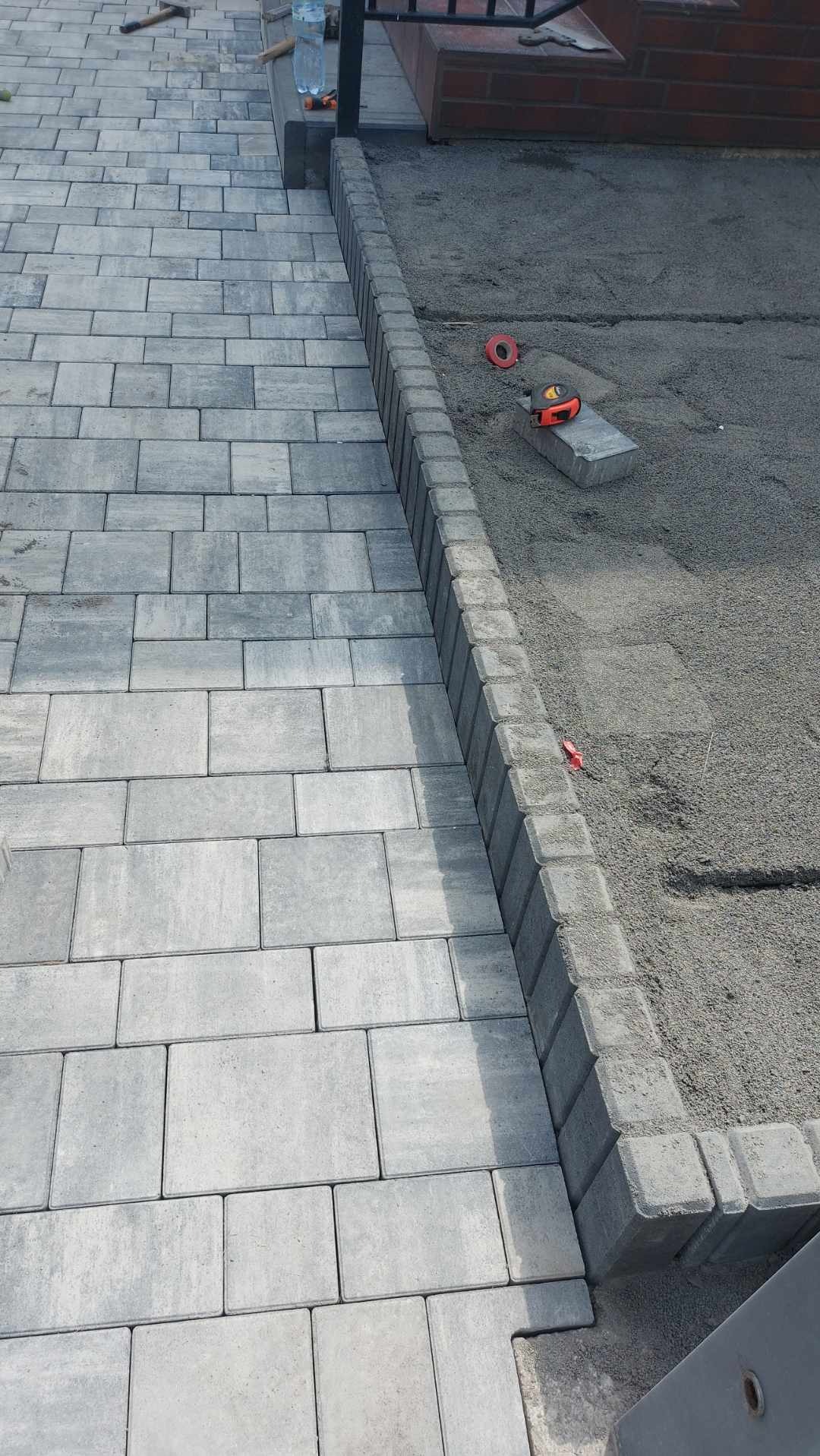 Fragment nawierzchni z szarej kostki brukowej, obrzeże z kostki, przygotowane podłoże z piasku, widoczna miarka i poziomica w tle.