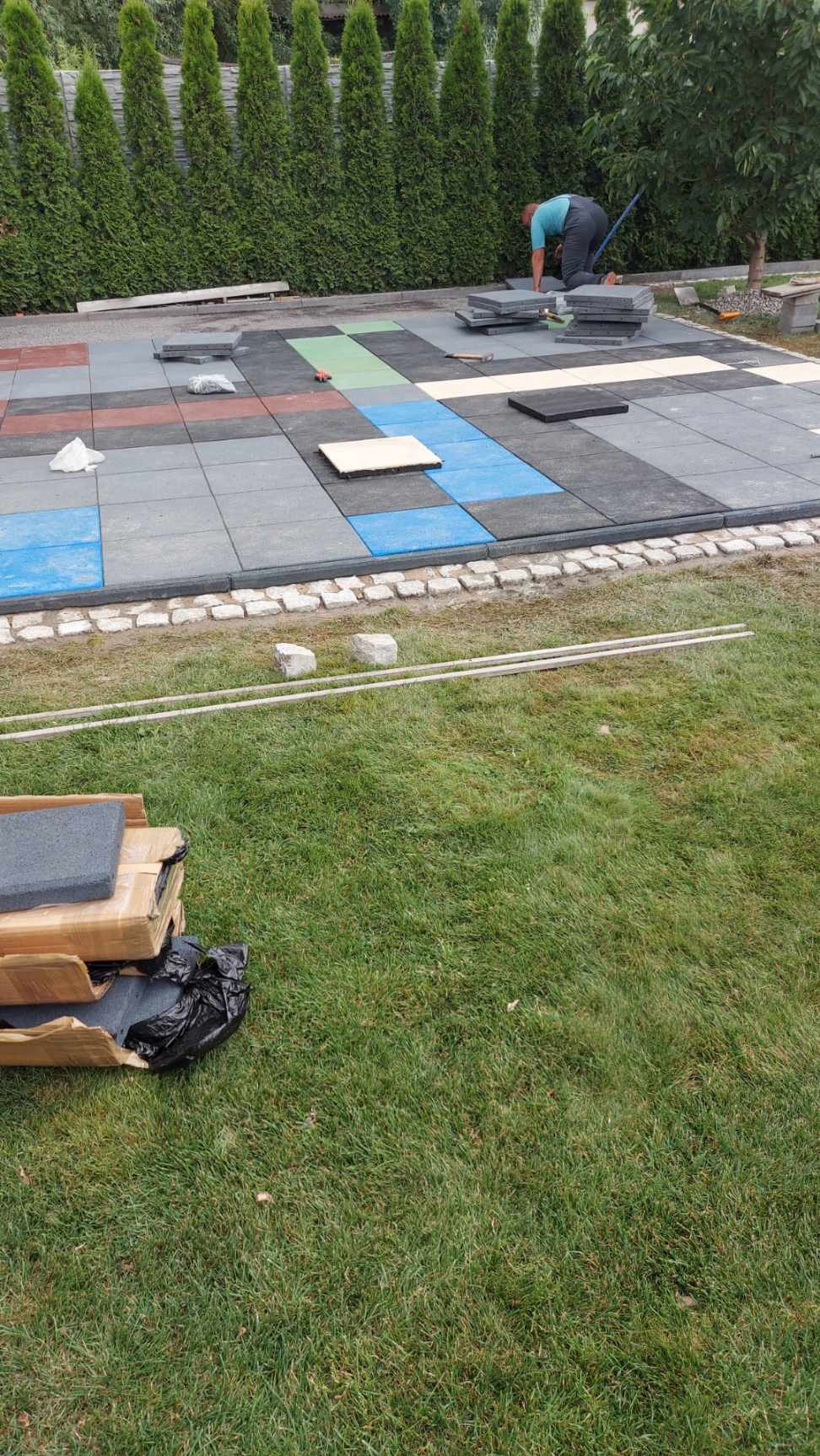 Pracownik układa kolorowe, gumowe płyty chodnikowe na trawniku, obok widoczne pakiety płyt i narzędzia.