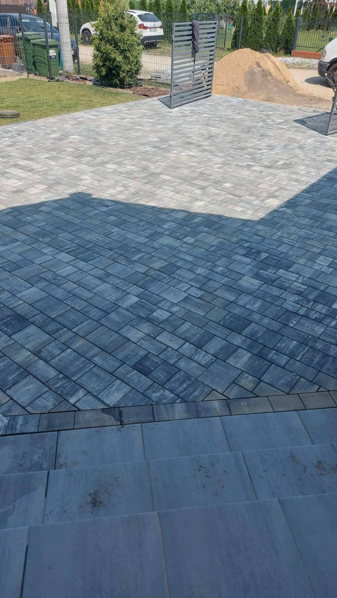 Ułożona kostka brukowa w szarych odcieniach, ułożona pod kątem, widoczne schody z kostki, w tle trawnik, drzewa i ogrodzenie.