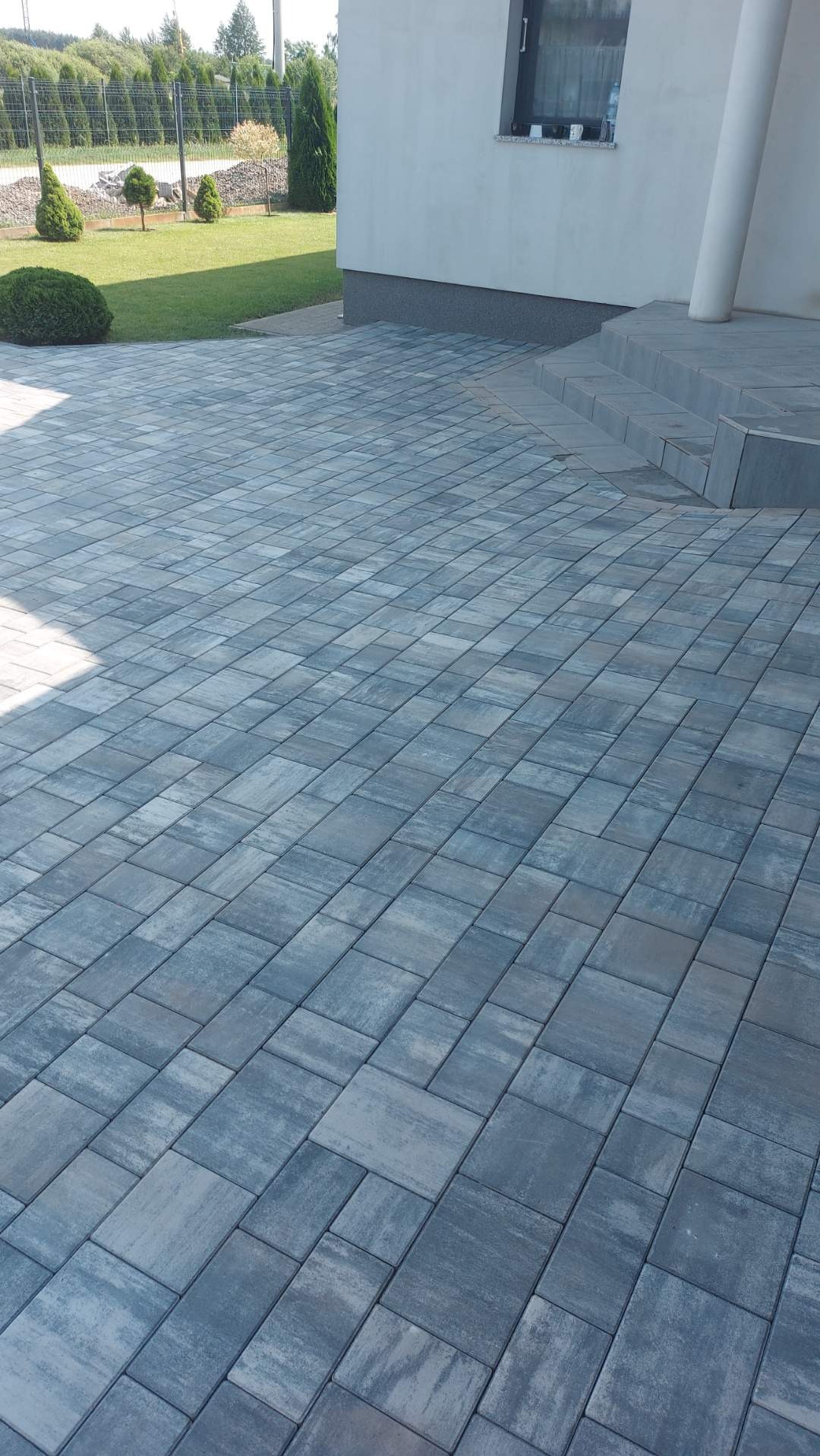 Nowo ułożona, szara kostka brukowa w jodełkę prowadząca do schodów wejściowych domu z kolumną, otoczona zielonym trawnikiem z ozdobnymi krzewami.
