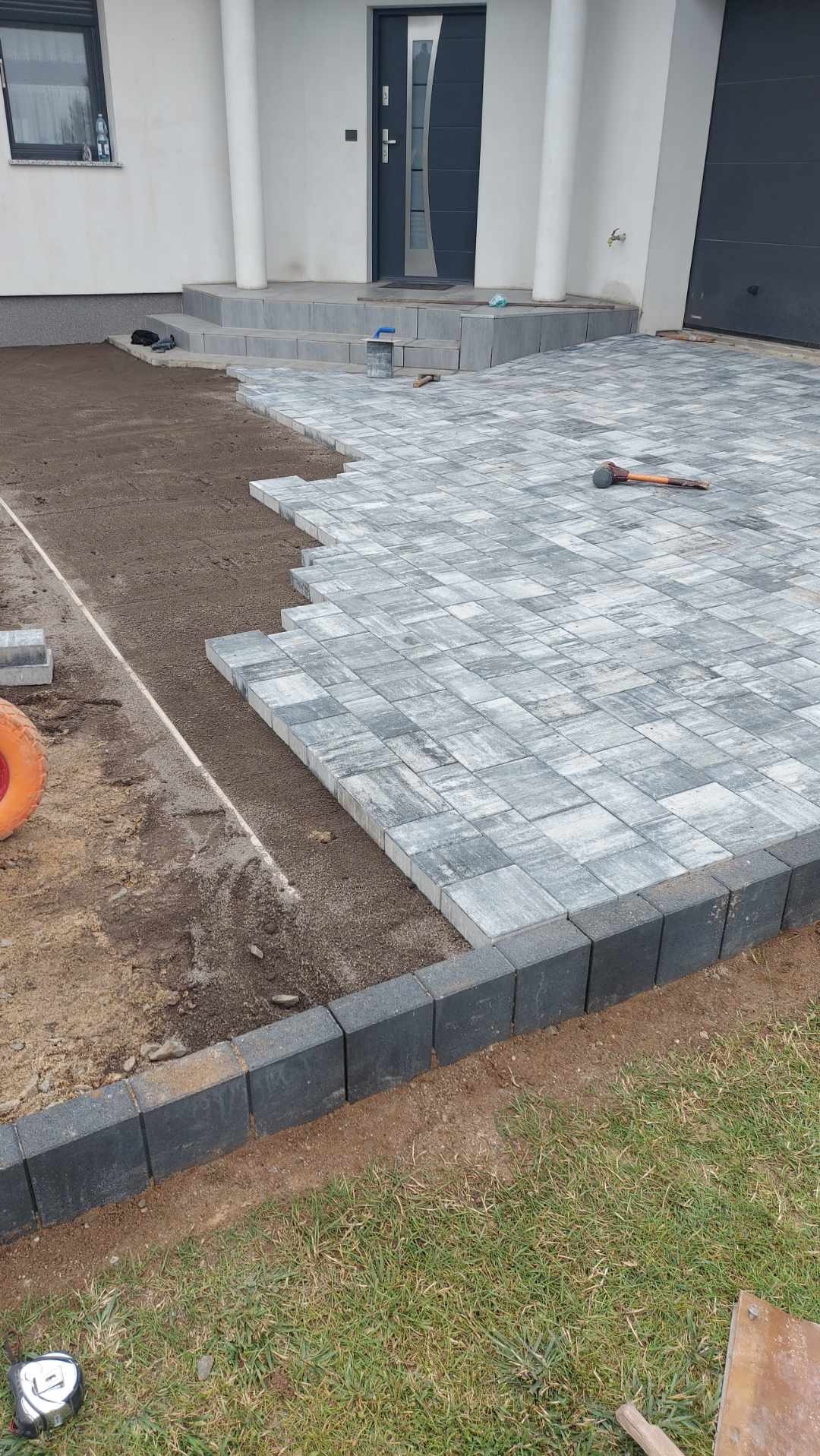 Niedokończona nawierzchnia z szarej kostki brukowej ułożonej w romby, widoczny gumowy młotek, obrzeża z ciemnej kostki, przygotowane podłoże i fragment trawnika.