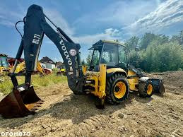 Żółta koparko-ładowarka Volvo BL71 na nierównym terenie, widok z boku, z łyżką skierowaną w dół, w tle drzewa i fragment budynku.