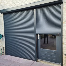 WOJCIECH FĄFARA BRAMSTEEL - Zamknięta, grafitowa roleta zewnętrzna z częściowo odsłoniętym oknem w drzwiach, odbijającym sylwetkę osoby na tle ceglanej ściany.