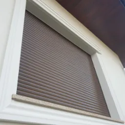 Zasłonięte okno z brązową roletą zewnętrzną, biała rama okienna z kamiennym parapetem, widoczny fragment drewnianego podbitki dachowej.