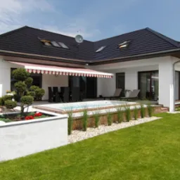 Biały dom z ciemnym dachem, tarasem i markizą w czerwono-białe paski, z zadbanym ogrodem i basenem.