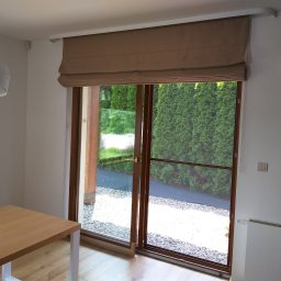 WOJCIECH FĄFARA BRAMSTEEL - Beżowa, rzymska roleta na oknie tarasowym w Raciechowicach, widok na ogród z zielonymi tujami i żwirowym podłożem, jasne wnętrze z drewnianym stołem.