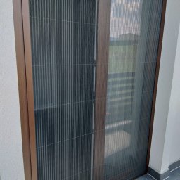 WOJCIECH FĄFARA BRAMSTEEL - Zewnętrzna plisa okienna w brązowej ramie, widok z balkonu, delikatne światło dzienne.