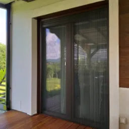 Zewnętrzna plisowana moskitiera w brązowej ramie, zamontowana na tarasie z drewnianą podłogą, widok na zieleń za oknem.