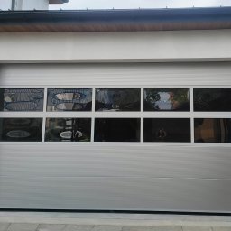 WOJCIECH FĄFARA BRAMSTEEL - Srebrna segmentowa brama z poziomymi panelami i rzędem okienek w górnej części, odbijających otoczenie.