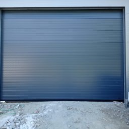 WOJCIECH FĄFARA BRAMSTEEL - Szara segmentowa konstrukcja zamknięta, zamontowana w hali przemysłowej, widoczne elementy konstrukcyjne budynku i gruz budowlany.