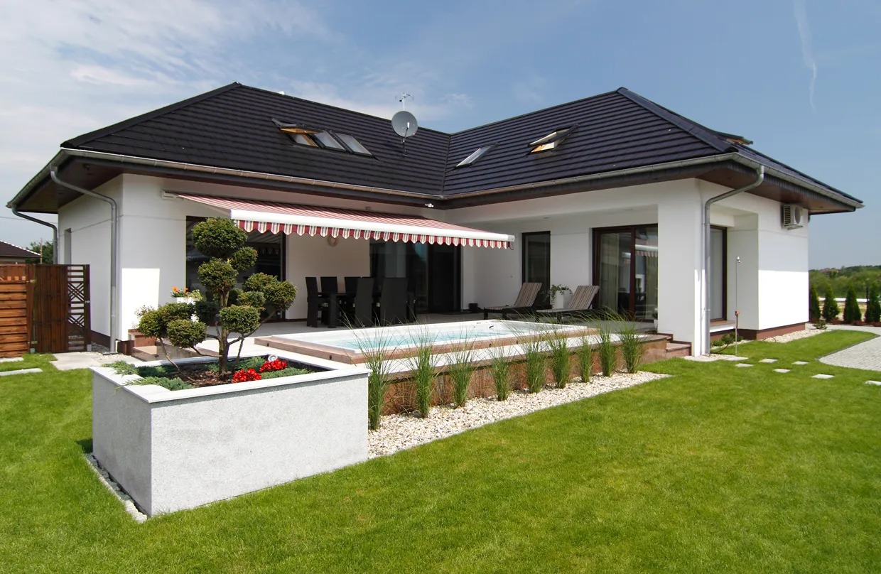 Biały dom z ciemnym dachem, tarasem i markizą w czerwono-białe paski, z zadbanym ogrodem i basenem.