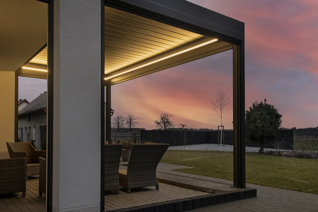 Nowoczesna pergola z oświetleniem LED, widok na ogród i zachodzące słońce, rattanowe krzesła wewnątrz.