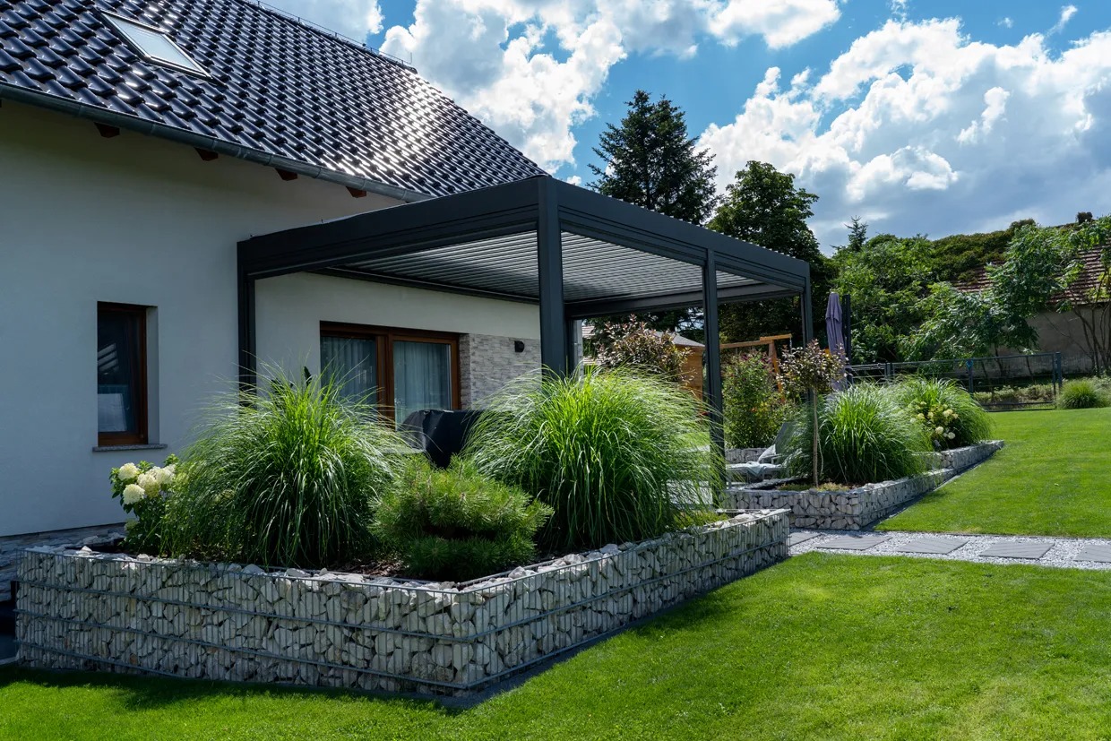 Nowoczesna, grafitowa konstrukcja zadaszenia tarasu z metalowymi lamelami, przylegająca do białego domu z ciemną dachówką, otoczona dekoracyjnymi rabatami z kamienia i ozdobnymi trawami na tle...