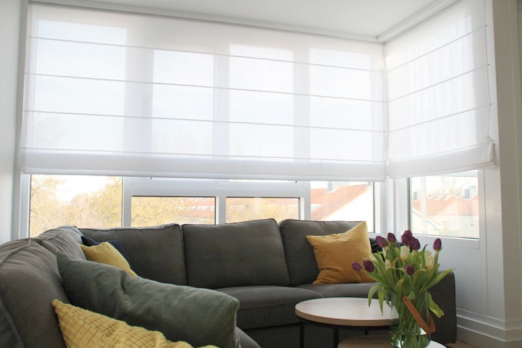 Jasne, białe, rzymskie osłony okienne zamontowane w rogu pokoju, częściowo zasłonięte przez szarą sofę z żółtymi poduszkami i tulipanami na stoliku kawowym.