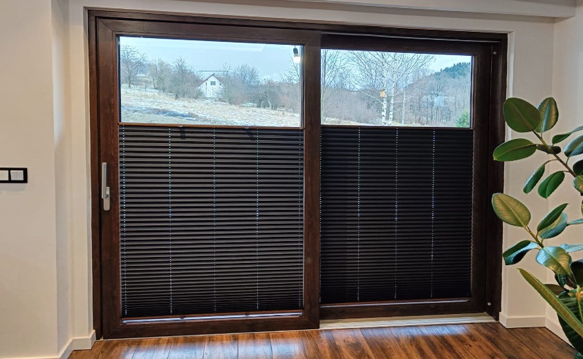 Ciemne, plisowane osłony okienne na dużym oknie przesuwnym z brązową ramą, widok na zimowy krajobraz z domem w tle, drewniana podłoga, roślina doniczkowa po prawej stronie.