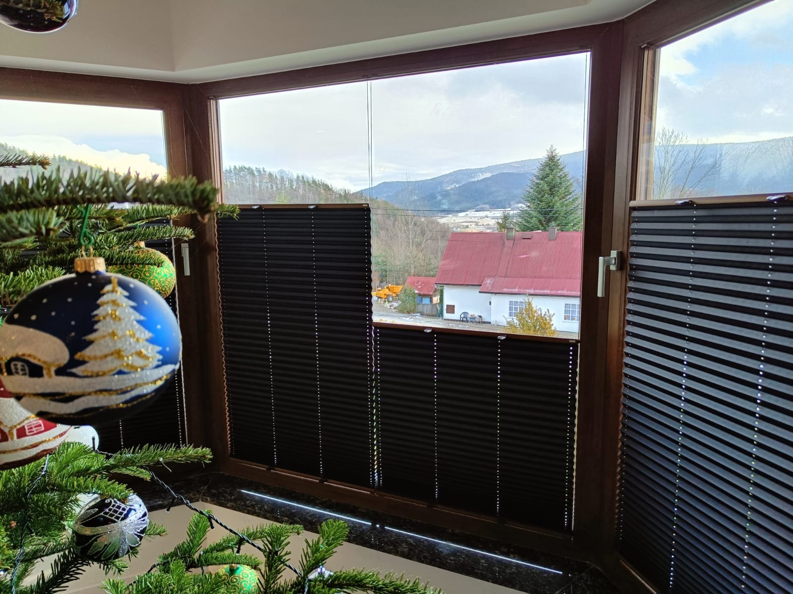 Ciemne, plisowane osłony okienne w brązowej ramie, częściowo zasłonięte, z widokiem na zimowy krajobraz górski i choinkę z ozdobami w pomieszczeniu.