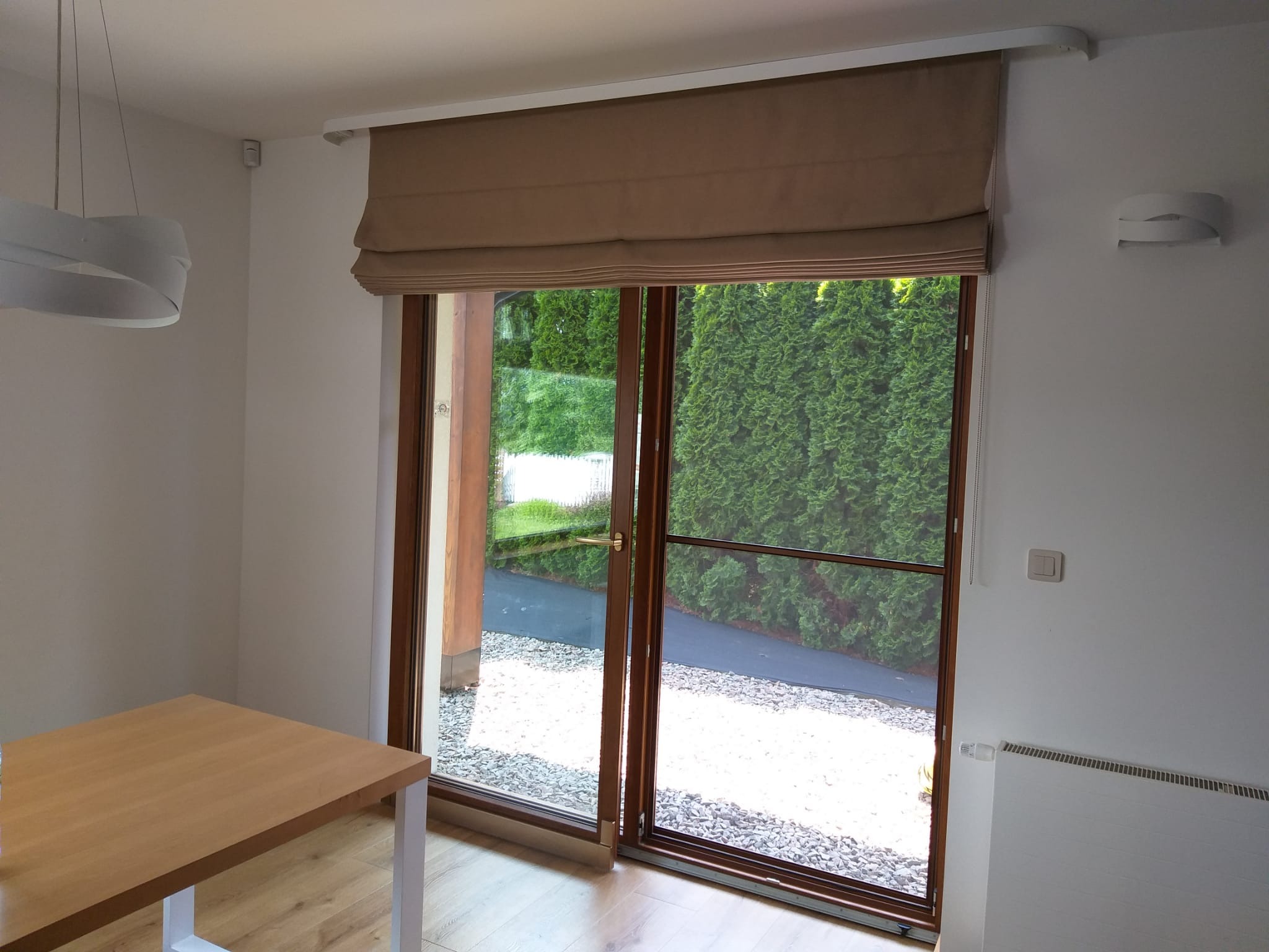 Beżowa, rzymska roleta na oknie tarasowym w Raciechowicach, widok na ogród z zielonymi tujami i żwirowym podłożem, jasne wnętrze z drewnianym stołem.