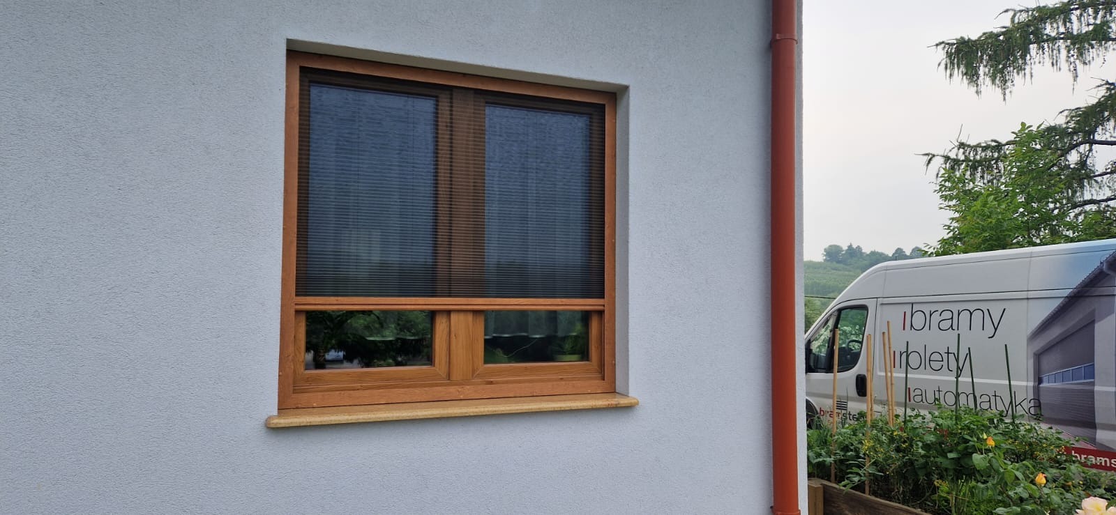 Okno z drewnianą ramą i plisowaną roletą wewnętrzną, widok z zewnątrz na tle jasnej elewacji budynku i zaparkowanego samochodu z reklamą firmy.