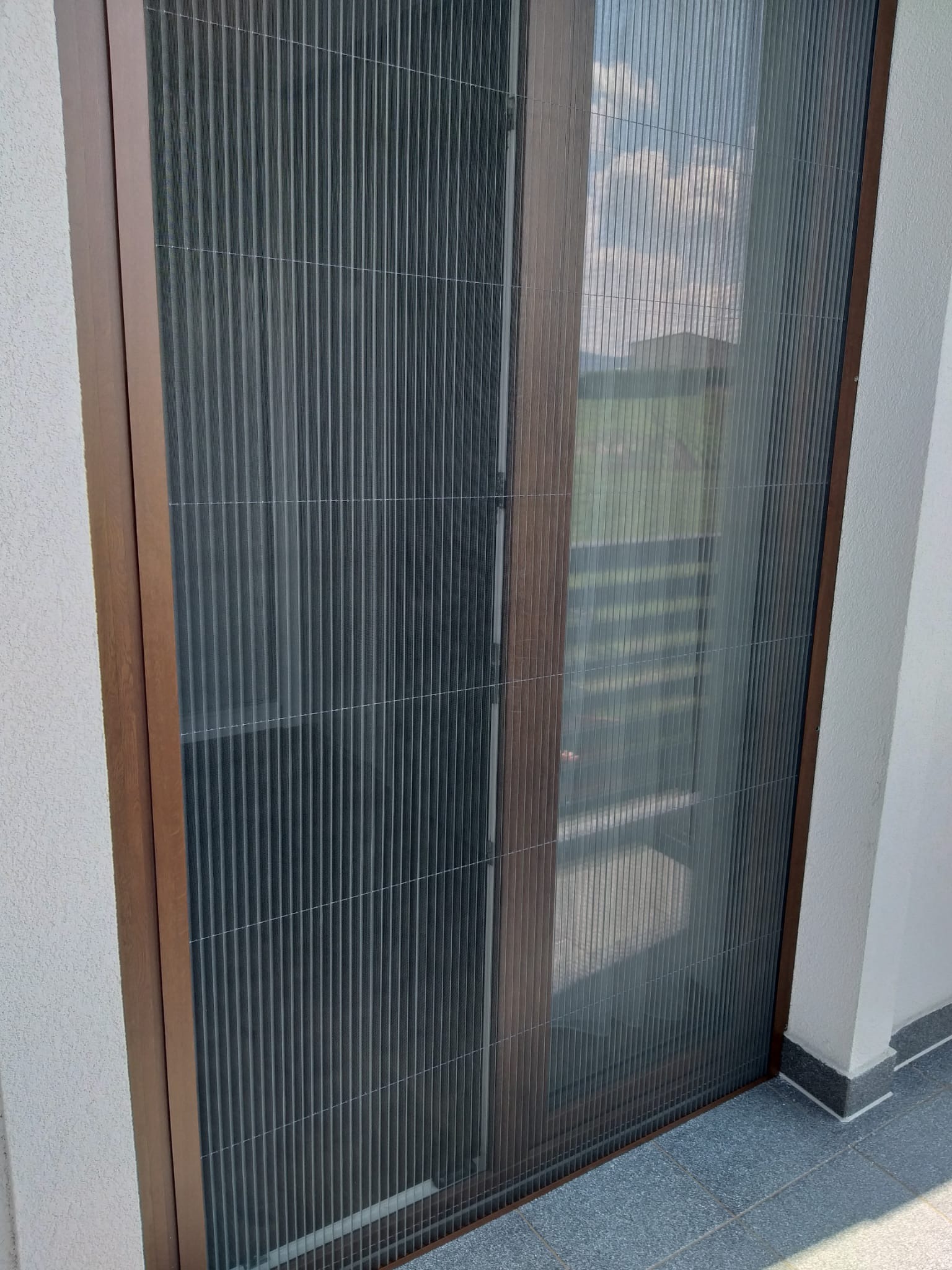 Zewnętrzna plisa okienna w brązowej ramie, widok z balkonu, delikatne światło dzienne.