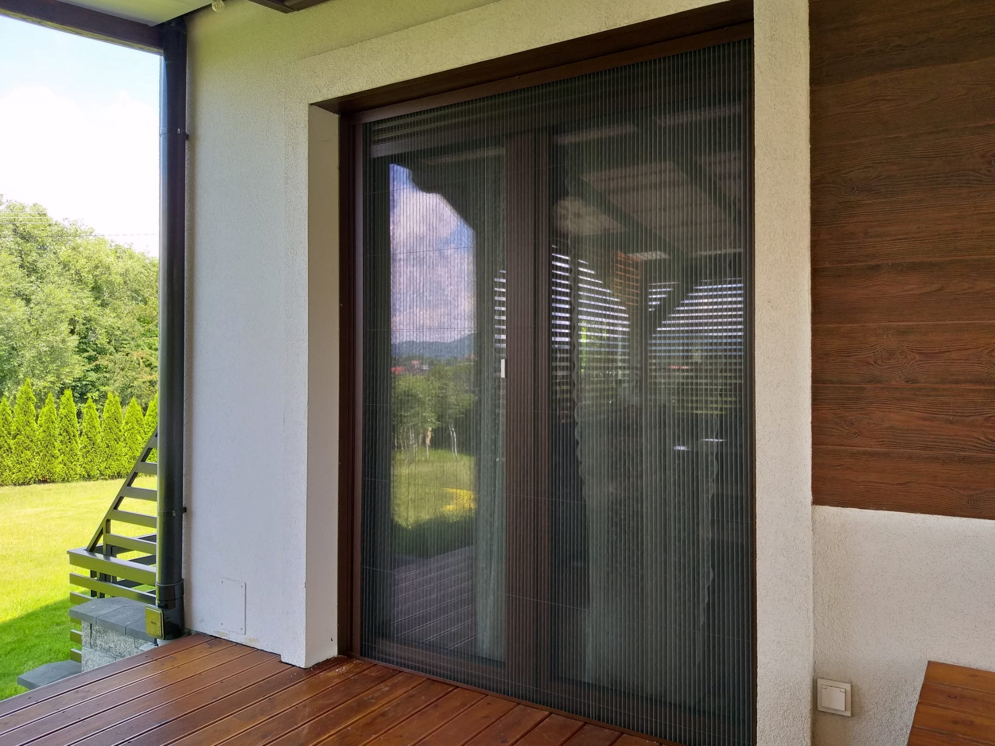 Zewnętrzna plisowana moskitiera w brązowej ramie, zamontowana na tarasie z drewnianą podłogą, widok na zieleń za oknem.